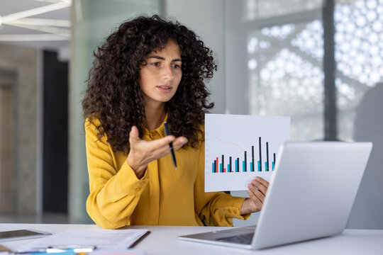 Hispanic businesswoman engaging in video call presentation, explaining data analysis while holding paper chart. Confident expression, professional setting, demonstrates expertise in business - Powered by Adobe