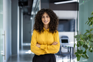 Hispanic businesswoman stands confidently with arms crossed in modern office. She smiles, conveying...