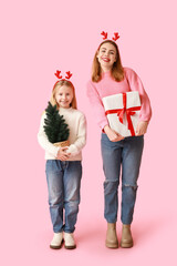 Cute little girl in reindeer horns with small Christmas tree and her mother holding gift box on pink background