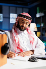 Detailed illustration of a focused Middle Eastern man in traditional attire writing on a notebook at a table. Close-up of an Arab man taking research notes with a pen in his home office.