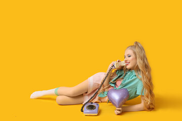 Fototapeta premium Young happy woman with retro telephone and heart-shaped balloon on yellow background