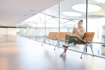 Young woman waits for news using her mobile phone sitting on a bench located in a passageway of a...