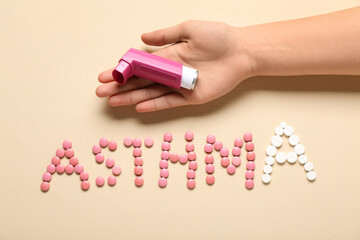 Child's hand with inhaler and word ASTHMA made of pills on beige background