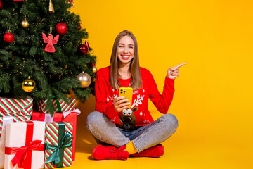 Full body portrait of nice young girl holdphone direct finger empty space wear festive x-mas new year sweater isolated on yellow color background