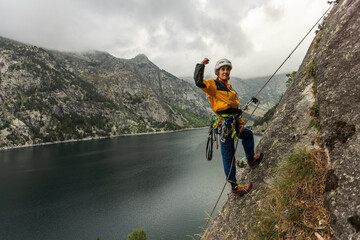 A man in a yellow jacket is standing on a rock near a body of water. He is wearing a helmet and he is enjoying the view