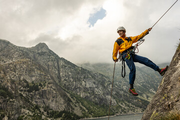 A man in a yellow jacket is climbing a mountain. He is wearing a helmet and harness. The mountain is covered in clouds, and the sky is overcast