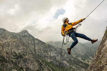 A man is jumping off a cliff with a rope attached to him. The man is wearing a yellow jacket and...