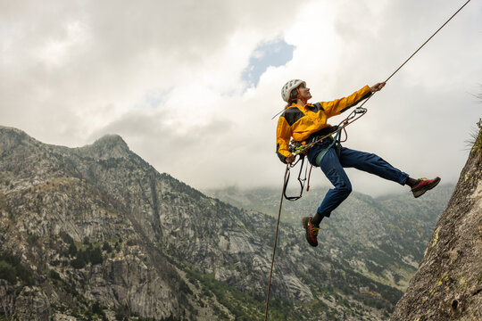 A man is jumping off a cliff with a rope attached to him. The man is wearing a yellow jacket and blue jeans. The sky is cloudy and the mountains are in the background. The man is wearing a helmet - Powered by Adobe