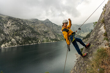 Obraz premium A woman is standing on a rock with a rope around her waist. She is wearing a yellow jacket and a helmet. The scene is set in a mountainous area with a lake in the background