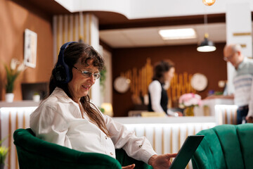 Detailed shot of old woman relaxing with headphones, using wireless computer in hotel foyer. Close up of elderly female tourist on sofa, listening to music and browsing the internet on laptop.