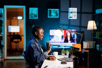 African american blogger waving at her personal computer while seated at a desk having a video call. Black woman uses digital laptop to chat online and research while immersed in digital technology.