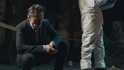 Detective inspecting area at crime scene at night as forensic experts collect evidence behind...