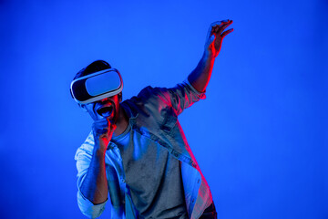 Excited man wearing VR goggle to exploring and enter in virtual program. Skilled gamer touching in metaverse while wearing casual cloth and standing at neon light background. Lifestyle. Deviation.