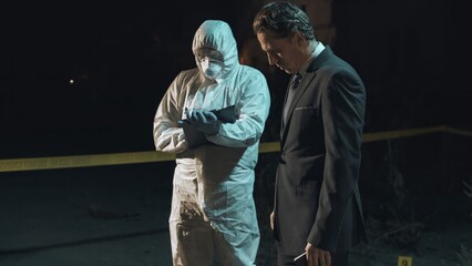Detective examining main clue brought in by forensic scientist at crime scene, while the rest of the experts collect remaining evidence behind police tape next to headless body. Night scene.