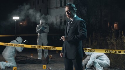 Detective inspecting area and smoking at crime scene at night as forensic experts collect evidence behind police tape next to headless body.