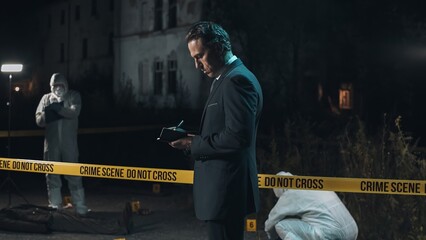 Detective inspecting area and taking notes in notebook at crime scene at night as forensic experts...