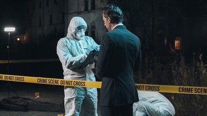 Detective inspecting area and taking notes in notebook at crime scene at night as forensic experts...