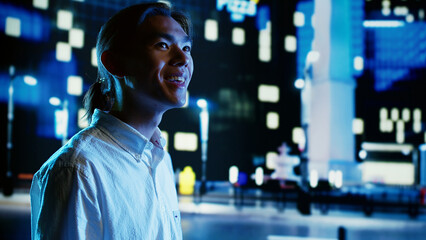 Asian man wandering around city avenues during nighttime, looking upwards to surrounding buildings...