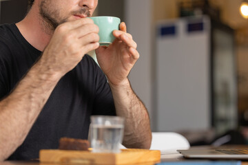 Freelance happy young man enjoying a delicious breakfast of latte and brouni in a specialty cafe, works with his laptop and smartphone, digital nomad, thoughtfully enjoys a cappuccino coffee