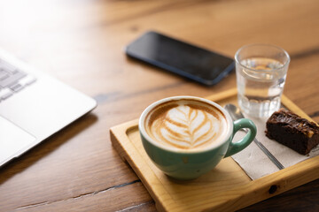 Close-up breakfast coffee with milk in the shape of a heart, latte art with pastries and glass of water, smartphone and laptop on a wooden table, energy for the whole day, espresso coffee