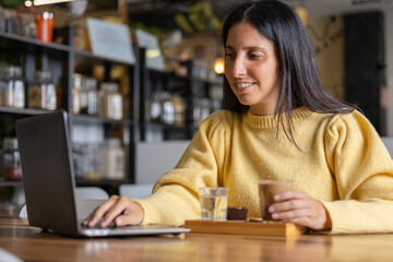 Freelance young happy woman enjoying a delicious breakfast of caramel coffee and brouni in a specialized cafe, works with her laptop, digital nomad, thoughtfully enjoys a coffee on a wooden table