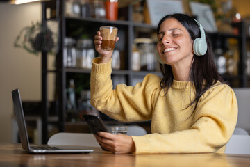 Freelance happy young woman enjoying a caramel and brownie coffee in a specialized coffee shop, works with her laptop, listening to music with headphones, digital nomad, enjoys a coffee