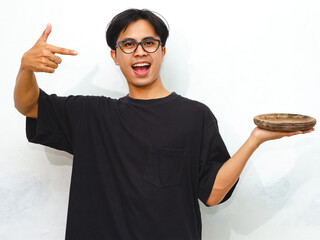 Portrait of an enthusiastic young Asian man wearing a blank t-shirt, pointing to an empty wooden...