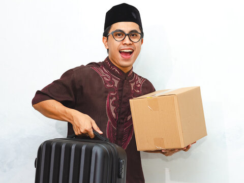 Cheerful Asian Muslim man carrying a suitcase and box, preparing for a flight to celebrate Eid Mubarak. Isolated on white background. Ideal for concepts of Ramadan and mudik travel.