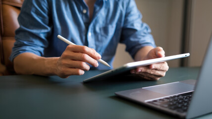 A man is writing on a tablet with a pen. He is sitting at a desk with a laptop in front of him