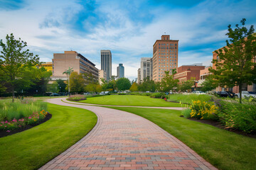 A curved brick road that leads to downtown is flanked on both sides by grass, flowers, and trees in the park's green space.