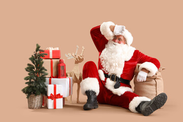 Tired Santa Claus with bag, Christmas tree and gift boxes sitting on beige background