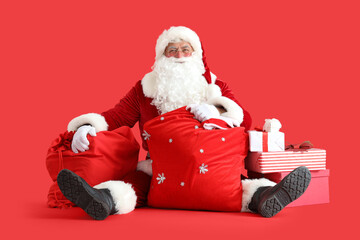 Santa Claus with bags and Christmas gift boxes sitting on red background