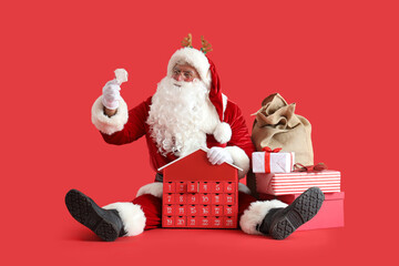 Santa Claus in deer horns with bag, gift boxes and Christmas advent calendar sitting on red background