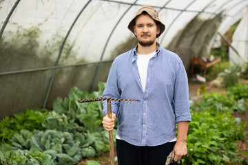 Man worker shirt is standing in greenhouse with rake and other tools in hands. He is resting after...