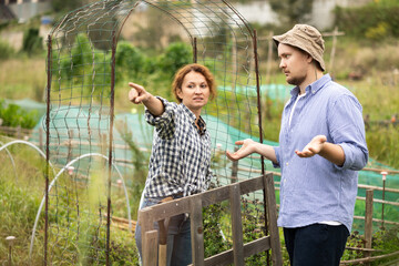 Female worker in jeans and checkered shirt is arguing with male neighbor of garden and country...