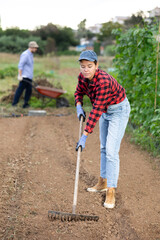 Peasant girl is working in garden plantation with rake. She levels and loosens soil with hand tool...