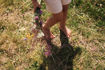 Fototapeta premium Little girl with bouquet of wildflowers walking barefoot outdoors on sunny day, closeup. Child enjoying beautiful nature