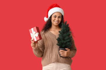 Happy African-American woman in Santa hat, with Christmas gift and fir tree on red background