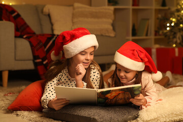 Cute happy little girls in Santa hats with book reading Christmas story at night