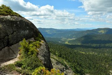 Obraz premium A rocky massif overgrown with grass on top of a mountain overlooking a dense coniferous forest in a picturesque valley under a cloudy summer sky.