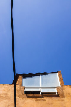 The photo captures a minimalist view of a building facade against a clear blue sky. A single, modern window sits within a rustic, textured stucco wall, partially open, allowing natural light to cast s