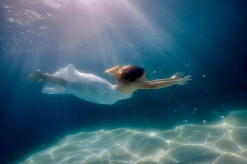 A girl in a white dress swims through a pool bathed in light rays from above