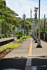 Naklejka premium A sunlit platform by the rail tracks features bright yellow tactile paving for the visually impaired, enhancing accessibility and adding contrast to the station scene.