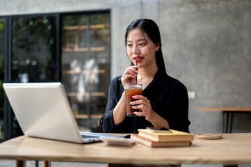 A beautiful Asian businesswoman enjoys iced coffee while working on her laptop, working remotely.