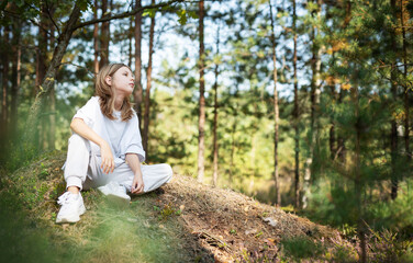 Young girl enjoying a sunny day in a forest clearing while sitting on a grassy mound surrounded by trees and nature