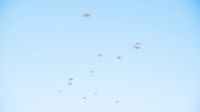 Group of parachutists in mid-descent form a coordinated pattern against a bright blue sky during a military skydiving exercise. The clean background enhances the visual appeal of their descent.