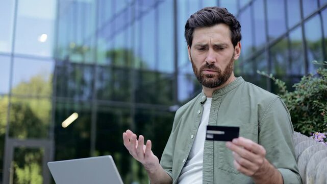 Concerned businessman holds credit card, struggling with online payment on laptop, experiencing stress and frustration. Business finance, online security, and technology issues concept.