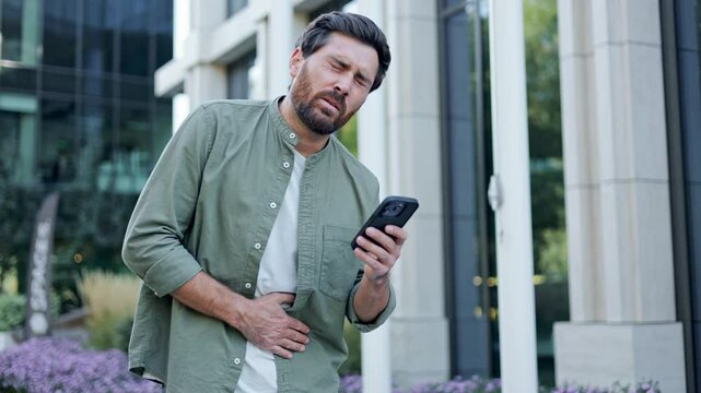 A concerned man experiencing stomach pain checks his phone for information or help. He's standing in an urban setting, highlighting the stress of city living and health anxieties.