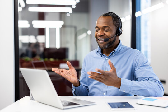 Smiling professional man using laptop for video conference call. Wearing headset, actively participating in meeting. Modern office setting suggests business environment with efficient communication.