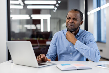 Man in blue shirt sitting at desk using laptop, displaying clear throat discomfort in modern office. Notebook and laptop on table create professional setting, conveying stress and health concerns